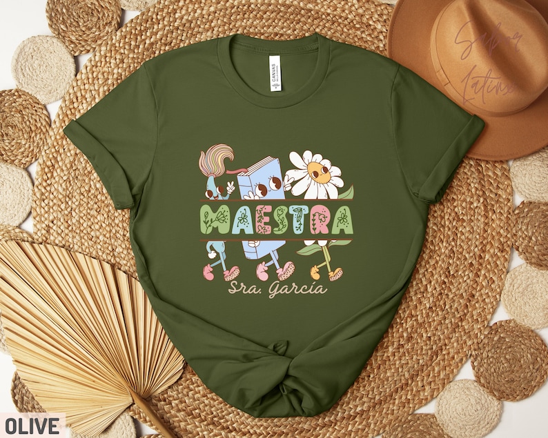 Peut inclure: T-shirt vert olive avec une illustration de dessin anim&eacute; d'un livre, d'une fleur et du mot "MAESTRA" avec le texte "Sra. Garcia" en dessous.