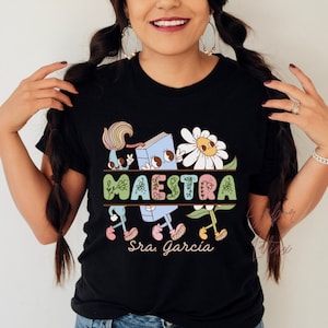 Peut inclure: T-shirt noir avec un motif graphique color&eacute; repr&eacute;sentant un livre, des fleurs et le mot "MAESTRA" avec le texte "Sra. Garcia" en dessous.
