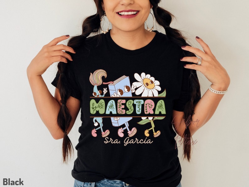 Peut inclure: T-shirt noir avec un motif graphique color&eacute; repr&eacute;sentant un livre, des fleurs et le mot "MAESTRA" avec le texte "Sra. Garcia" en dessous.