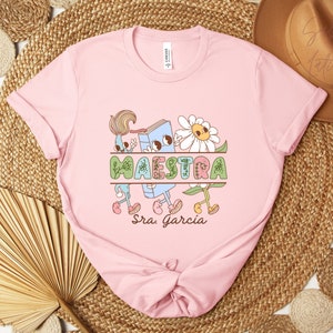 Peut inclure: T-shirt rose avec un graphique d'un livre, des fleurs et le mot "MAESTRA" avec le nom "Sra. Garcia" en dessous.