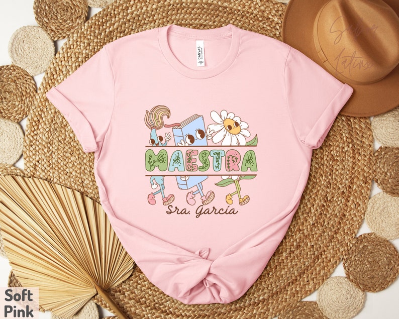 Peut inclure: T-shirt rose avec un graphique d'un livre, des fleurs et le mot "MAESTRA" avec le nom "Sra. Garcia" en dessous.