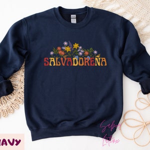 May include: Navy blue sweatshirt with a floral design and the text "SALVADORENA" in rainbow colors.