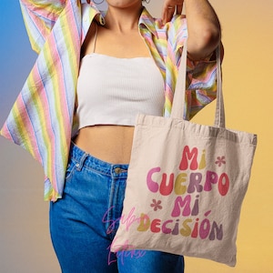 May include: Beige tote bag with the text "Mi Cuerpo Mi Decisi&oacute;n" in colorful, retro-style lettering. The bag has long handles and is made of a natural-colored fabric. The background is a gradient of yellow and blue.