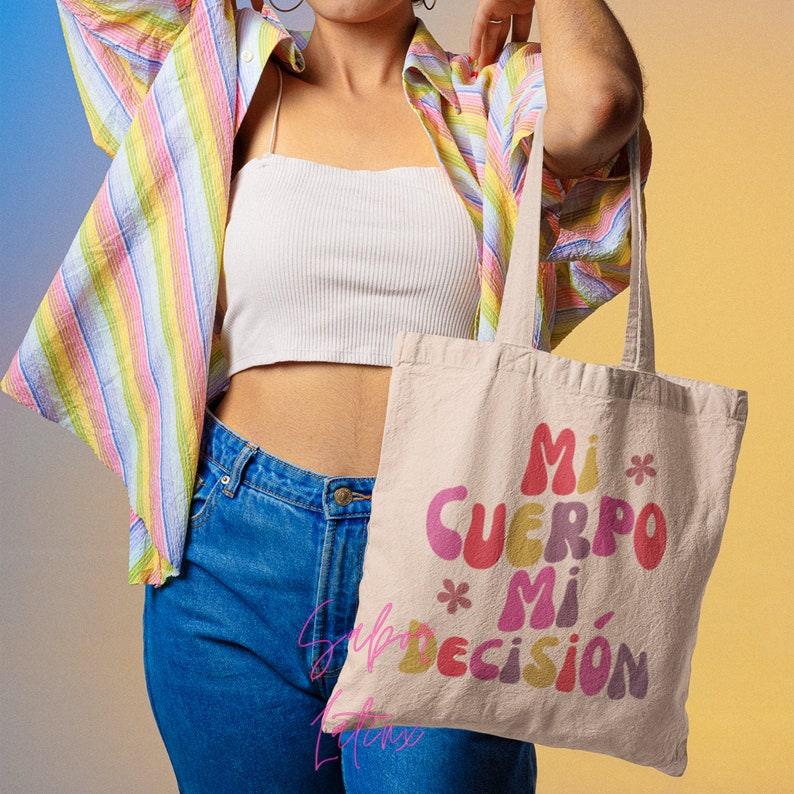 May include: Beige tote bag with the text "Mi Cuerpo Mi Decisi&oacute;n" in colorful, retro-style lettering. The bag has long handles and is made of a natural-colored fabric. The background is a gradient of yellow and blue.