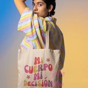May include: Beige canvas tote bag with the text "Mi Cuerpo Mi Decisi&oacute;n" in colorful, retro-style lettering. The bag has a simple design with two handles and is held by a person wearing a striped, pastel-colored shirt.