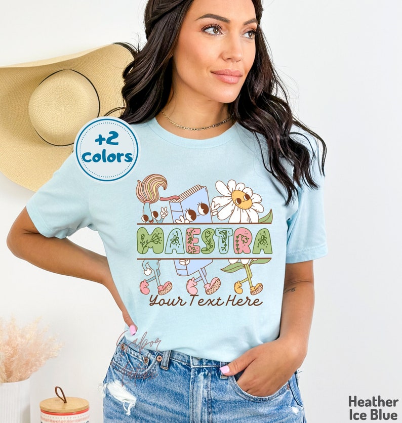 Peut inclure: Un t-shirt bleu clair avec un motif graphique repr&eacute;sentant un livre, une fleur et un pinceau. Le mot "MAESTRA" est &eacute;crit dans le motif. Le texte "Your Text Here" est situ&eacute; sous le graphique.