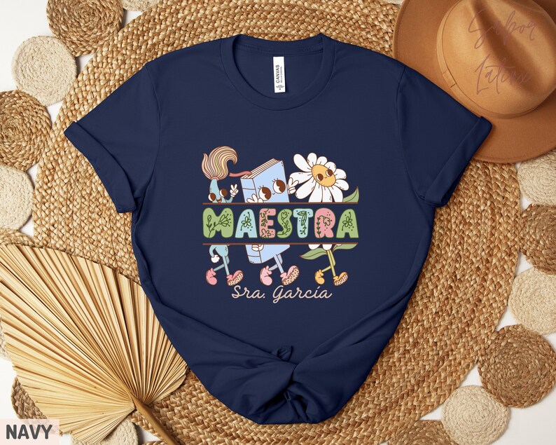 Peut inclure: Un t-shirt bleu marine avec un graphique repr&eacute;sentant un livre, des fleurs et le mot "MAESTRA" avec le texte "Sra. Garcia" en dessous.