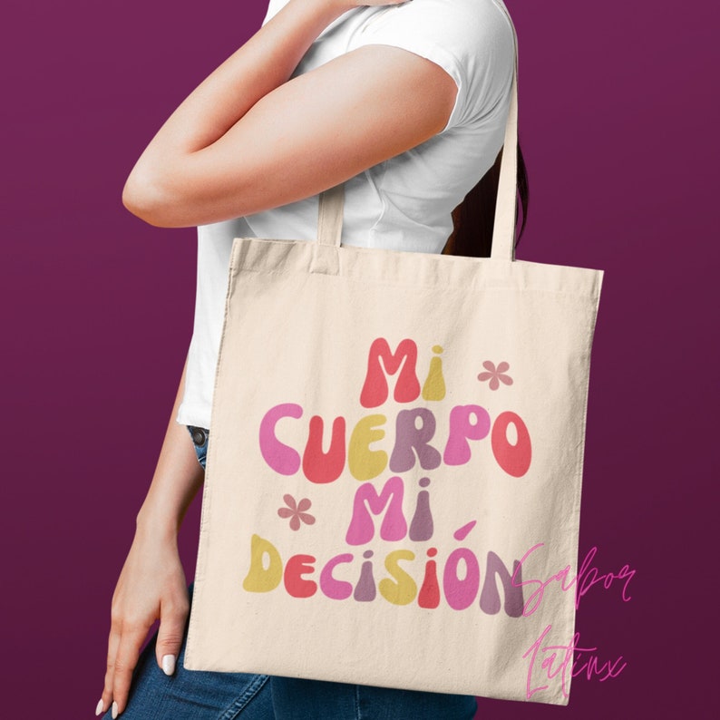May include: Cream-colored tote bag with the text "Mi Cuerpo Mi Decisi&oacute;n" in colorful, bubble-style letters. The bag has a simple design with a rectangular shape and long straps. The text is in shades of pink, yellow, and red.