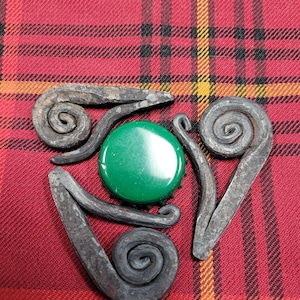May include: A metal triquetra design with three spirals, each with a different orientation. The triquetra is centered on a green bottle cap.