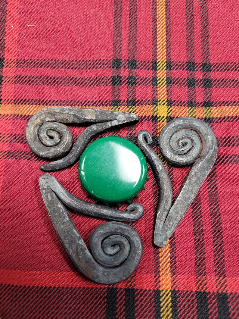 May include: A metal triquetra design with three spirals, each with a different orientation. The triquetra is centered on a green bottle cap.