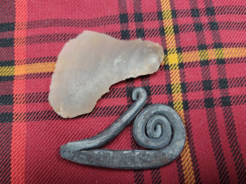 May include: A gray flint stone and a metal fire starter with a spiral design on a red and black plaid fabric background.