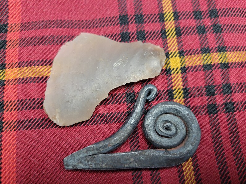 May include: A gray stone and a metal snail-shaped fire starter on a red and black plaid fabric.