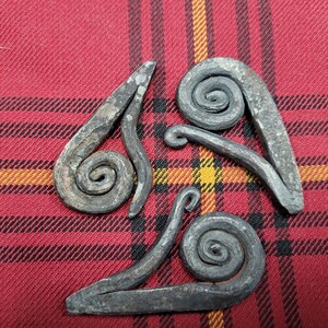 May include: Three metal pieces with a spiral design, arranged in a triangular shape on a red and black plaid fabric.