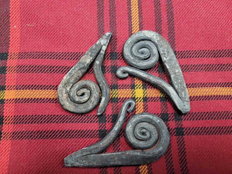 May include: Three metal pieces with a spiral design, arranged in a triangular shape on a red and black plaid fabric.