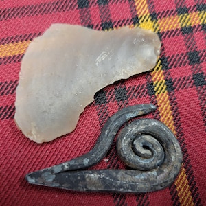May include: A light brown, irregularly shaped stone and a dark gray metal piece with a spiral design. The objects are on a red and black plaid fabric.