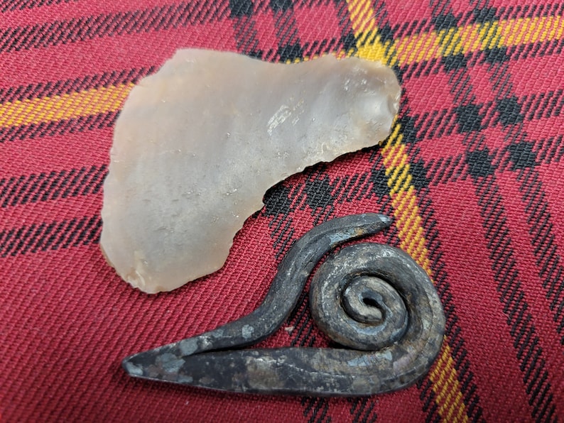 May include: A light brown, irregularly shaped stone and a dark gray metal piece with a spiral design. The objects are on a red and black plaid fabric.