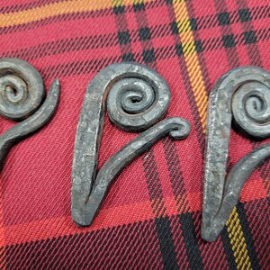 May include: Three metal swirls, each with a unique shape and design, are displayed on a red and yellow plaid fabric. The swirls are made of iron and have a rustic, hand-forged look.