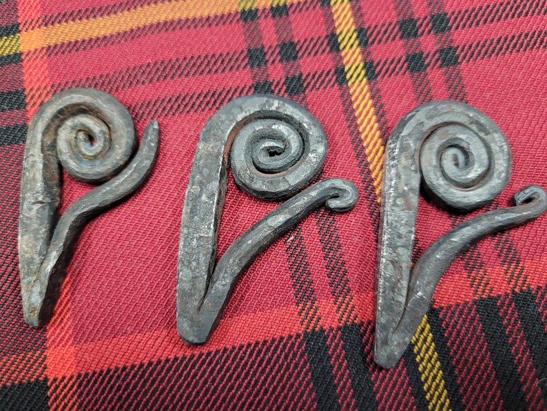 May include: Three metal swirls, each with a unique shape and design, are displayed on a red and yellow plaid fabric. The swirls are made of iron and have a rustic, hand-forged look.