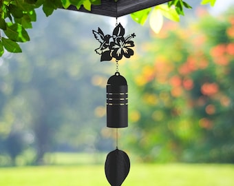 May include: Black metal wind chime featuring a hummingbird and hibiscus flower design. The chime has a cylindrical body with horizontal stripes and a leaf-shaped bottom. The wind chime hangs from a wooden structure, with a blurred green and orange background.