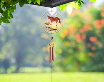 May include: A copper-colored wind chime featuring a circular design with a horse and foal silhouette. The chime hangs from a wooden structure, with a blurred green and orange background. The chime has several copper-colored tubes.