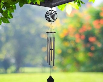 May include: Black metal wind chimes with a butterfly and floral design. The chimes hang from a wooden structure, with a blurred green and orange background. The wind chimes have multiple tubes and a diamond-shaped clapper.