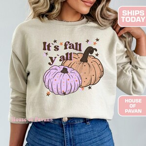 Può includere: Una felpa beige con il testo "It's fall y'all" e una grafica di due zucche, una rosa con pois bianchi e una arancione con macchie marroni.