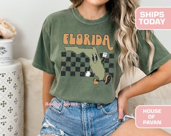May include: Olive green t-shirt featuring a cartoon Florida state graphic with the word "FLORIDA" in orange. The graphic includes a checkered background. The shirt is a comfortable, casual style.