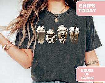 May include: A dark gray t-shirt with a graphic of four different coffee drinks. The drinks are a iced coffee, a latte, a coffee with whipped cream and a coffee with a cow print design. The text "House of Pavan" is on the shirt.
