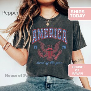 May include: A dark grey t-shirt with a vintage-style graphic of an eagle and the word "AMERICA" in blue and red lettering. The graphic also includes the numbers "17" and "76" and the text "land of the free".