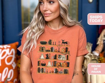 Puede incluir: Una mujer lleva una camiseta naranja quemada con un gráfico de una estantería llena de artículos espeluznantes con temática de Halloween. La camiseta tiene cuello redondo y mangas cortas. La mujer lleva una boina beige y pantalones cortos de mezclilla azul.
