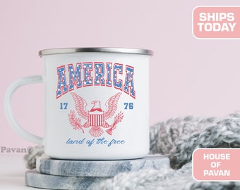 May include: White enamel mug with a red, white, and blue American flag design. The mug has a handle and says "AMERICA 17 76 land of the free".
