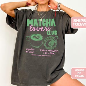 May include: Dark gray t-shirt with the text "Matcha Lovers Club" in green and pink. Below the text are illustrations of a matcha drink and whisk. The shirt also includes the text "regular or iced" and "every morning 7 am-11am".