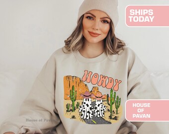 May include: A beige sweatshirt featuring a vintage-style graphic print of two cartoon cowboys riding horses in the desert. The graphic includes the text "HOWDY" in a colourful font.