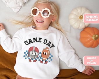 May include: White sweatshirt featuring a graphic of a football helmet and football with the text "GAME DAY" printed on it. The sweatshirt is being worn by a child with blonde hair and flower-shaped sunglasses.