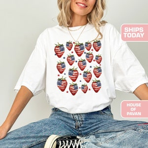 May include: White t-shirt with a pattern of strawberries designed as the American flag. The strawberries are red with blue and white details. Small red and blue stars are scattered around the strawberries. The text "SHIPS TODAY" is visible.