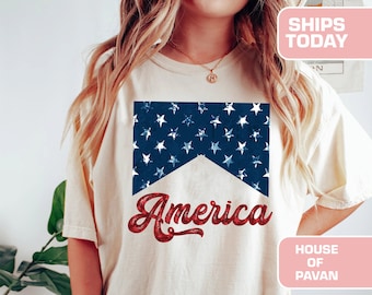 May include: A white t-shirt with a red, white, and blue American flag design and the word "America" in red glitter lettering. The shirt is being worn by a person. The text "House of Pavan" is visible in the bottom right corner of the image.