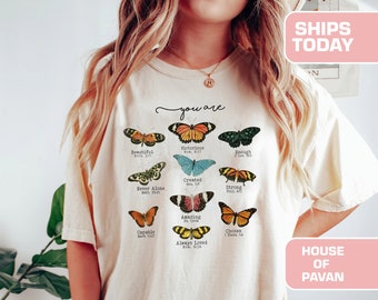 May include: A cream-coloured t-shirt with a butterfly design and the words "you are" above the butterflies. Each butterfly is a different colour and is labelled with a positive affirmation and a Bible verse reference. The shirt has short sleeves.
