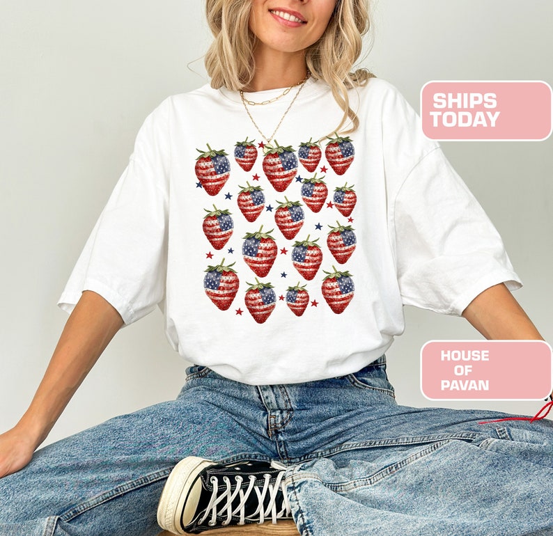 May include: White t-shirt with a pattern of strawberries designed as the American flag. The strawberries are red with blue and white details. Small red and blue stars are scattered around the strawberries. The text "SHIPS TODAY" is visible.