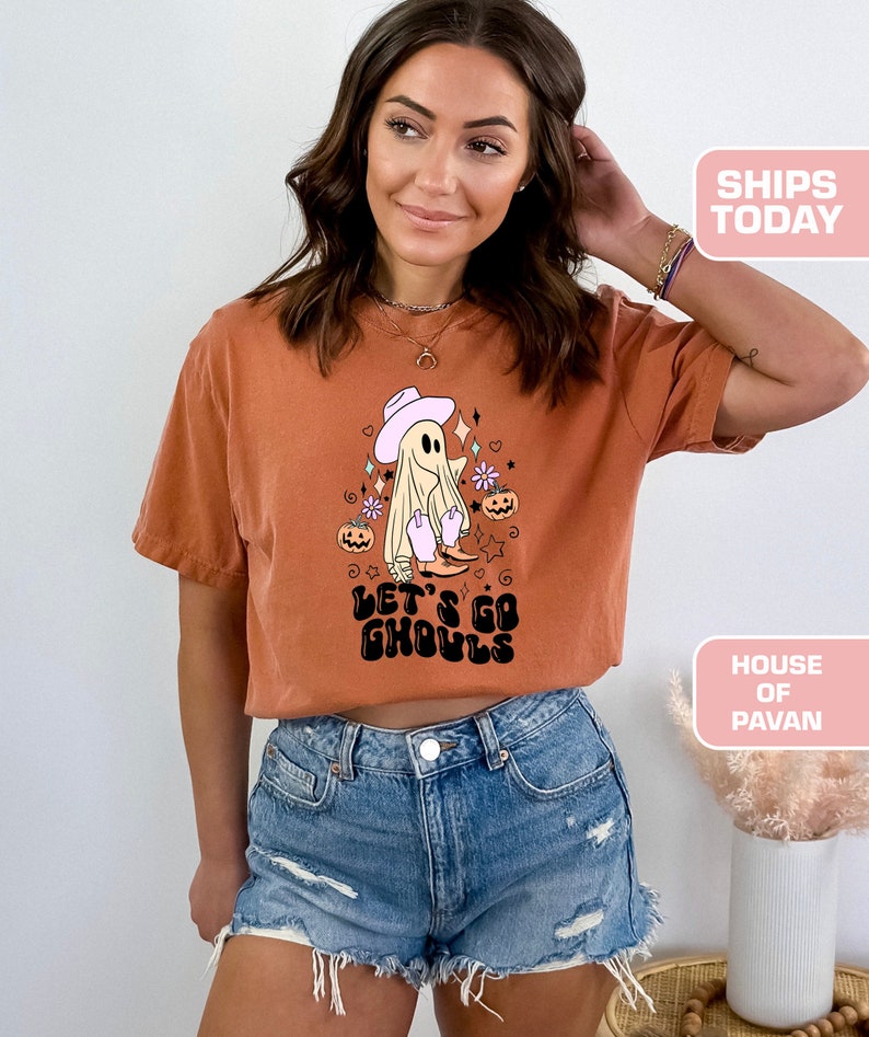 K&ouml;nnte beinhalten: Ein rostfarbenes T-Shirt mit einem Aufdruck eines Geists, der einen Cowboyhut und Stiefel tr&auml;gt. Der Text "Let's Go Ghouls" ist in Schwarz unter dem Geist gedruckt.