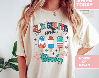 May include: A white t-shirt with a red, white and blue graphic design. The graphic features the text "Red White and Cool" with three popsicles decorated with the American flag. The popsicles are surrounded by stars and fireworks.