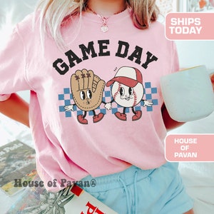 以下が含まれることがあります： 「GAME DAY」という文字がプリントされた、野球ボールと野球グローブのグラフィックが描かれたピンクのTシャツです。