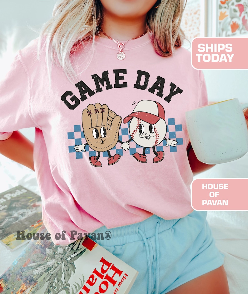 以下が含まれることがあります： 「GAME DAY」という文字がプリントされた、野球ボールと野球グローブのグラフィックが描かれたピンクのTシャツです。