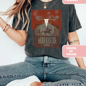 May include: A woman wearing a grey t-shirt with a vintage-style graphic of a skull with horns and a cowboy riding a horse. The graphic says "American Cowboy" and "Rodeo Show".