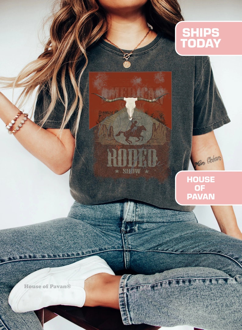 May include: A woman wearing a grey t-shirt with a vintage-style graphic of a skull with horns and a cowboy riding a horse. The graphic says "American Cowboy" and "Rodeo Show".