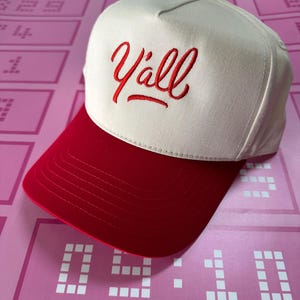 May include: A baseball cap with a cream-coloured crown and a red brim. The word "Y'all" is embroidered in red script on the front. The cap has a red button on top.