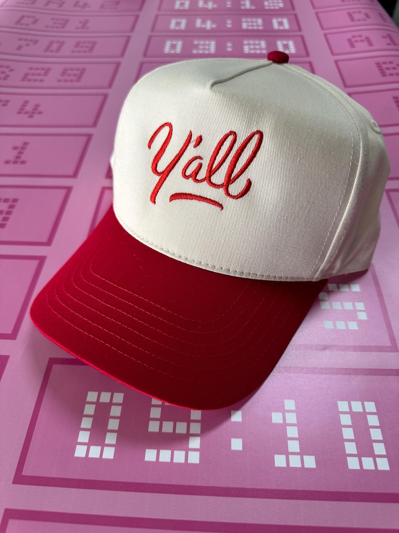 May include: A baseball cap with a cream-coloured crown and a red brim. The word "Y'all" is embroidered in red script on the front. The cap has a red button on top.