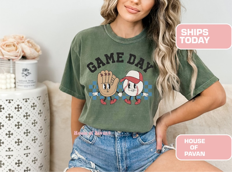 以下が含まれることがあります： 「GAME DAY」という文字がプリントされた、野球ボールとグローブのヴィンテージスタイルのグラフィックが描かれた緑色のTシャツです。