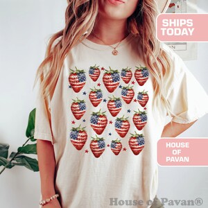 May include: Cream-coloured t-shirt featuring a pattern of strawberries with the American flag design. The strawberries are red and white with blue accents and green stems. The shirt has the text "House of Pavan".