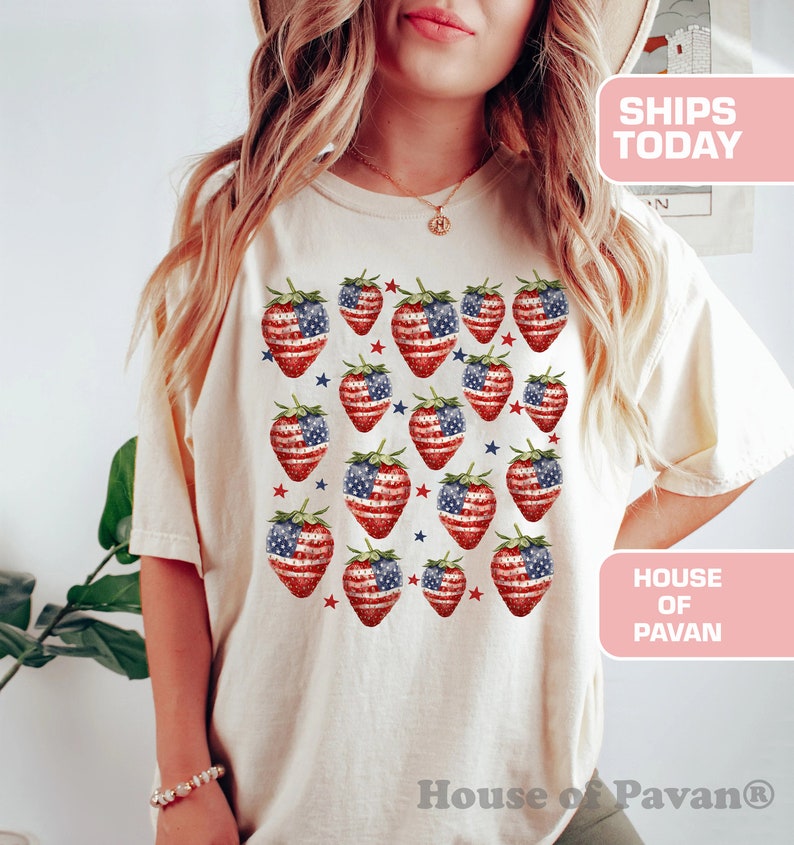 May include: Cream-coloured t-shirt featuring a pattern of strawberries with the American flag design. The strawberries are red and white with blue accents and green stems. The shirt has the text "House of Pavan".