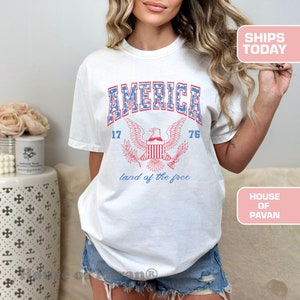 May include: White t-shirt with a vintage-style graphic of an eagle with the words "AMERICA 17 76 land of the free" printed on it.